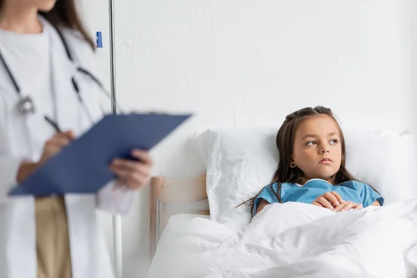 Грустный ребенок смотрит в сторону, лежа на кровати рядом с размытым доктором с планшетом — стоковое фото