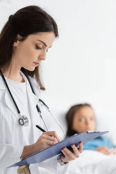 Pediatrician in white coat writing on clipboard near blurred kid in hospital — Stock Photo