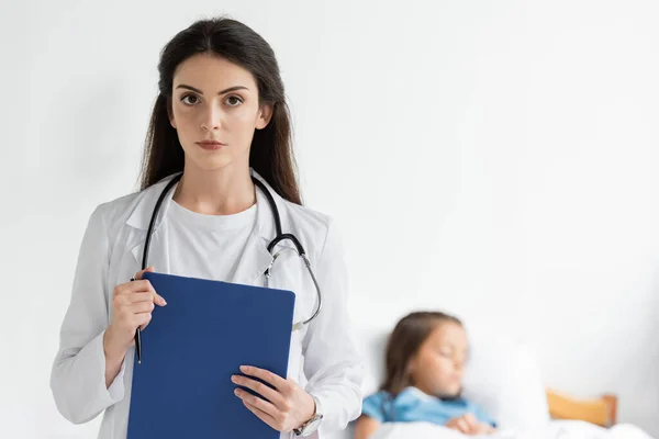 Pediatrician holding clipboard and looking at camera near blurred child in clinic — Stock Photo