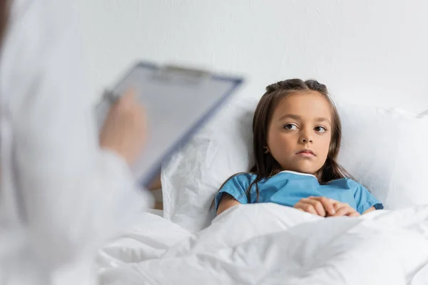 Kind liegt auf Klinikbett neben verschwommenem Arzt und schreibt auf Klemmbrett — Stockfoto