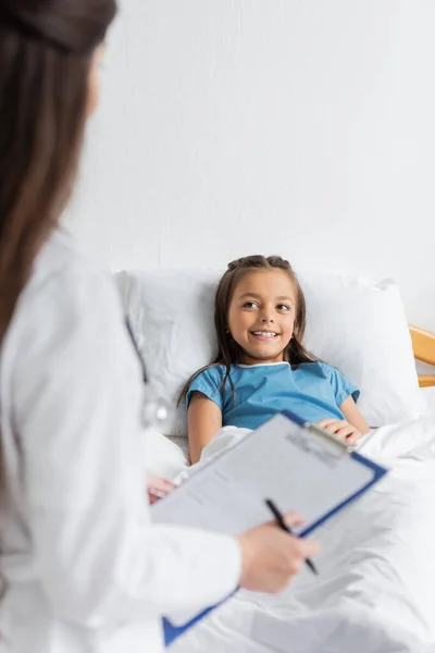 Smiling kid lying on bed while blurred doctor holding clipboard in hospital ward — Stock Photo