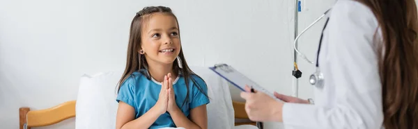 Chica haciendo por favor gesto cerca de pediatra con portapapeles en la clínica, pancarta - foto de stock