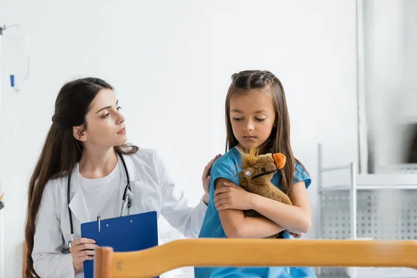 Pediatra sujetando portapapeles y calmando niño molesto con juguete suave en la sala de hospital - foto de stock