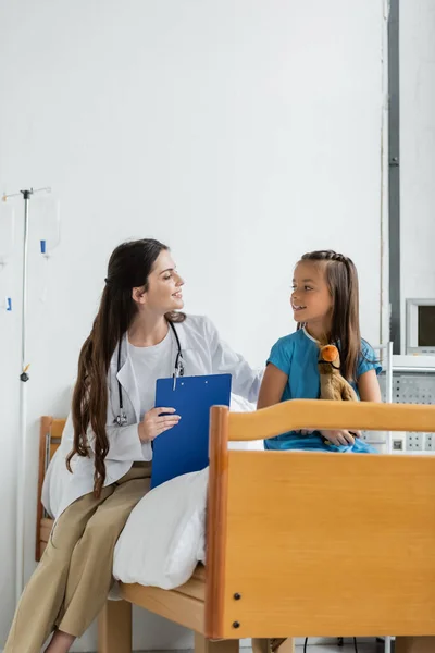 Smiling doctor holding clipboard and talking to child with toy on bed in clinic — Stock Photo