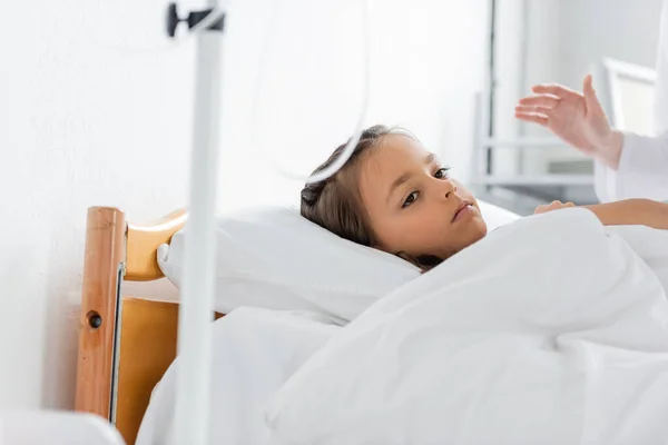 Niño triste acostado en la cama cerca del médico en la sala de hospital - foto de stock