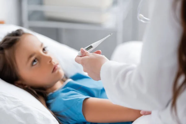 Doctor en bata blanca sosteniendo termómetro electrónico cerca de un niño borroso en la sala del hospital - foto de stock