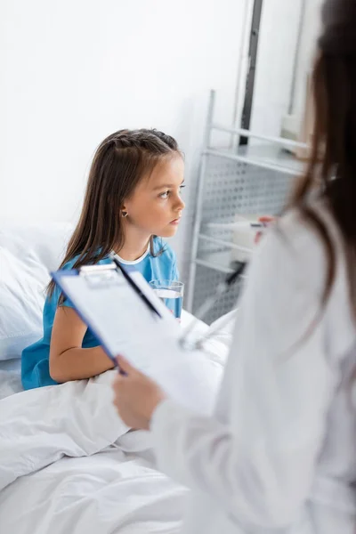 Child holding glass of water on bed near blurred pediatrician in hospital ward — Stock Photo