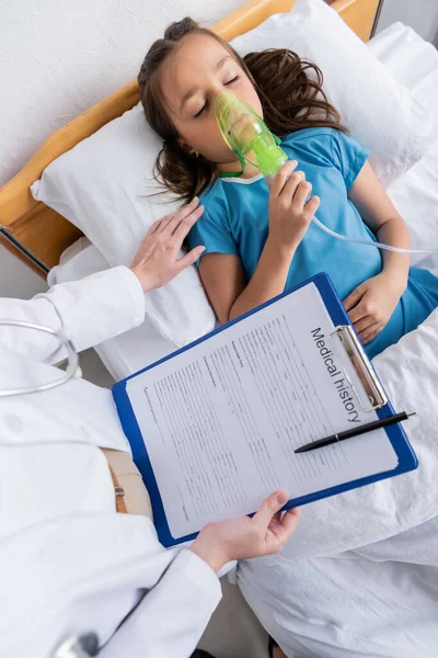 Top view of pediatrician holding clipboard near child with oxygen mask on bed in clinic — Stock Photo