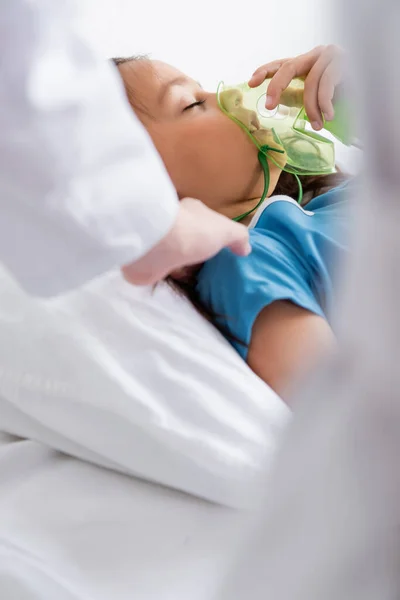 Blurred pediatrician touching child with oxygen mask on bed in clinic — Stock Photo