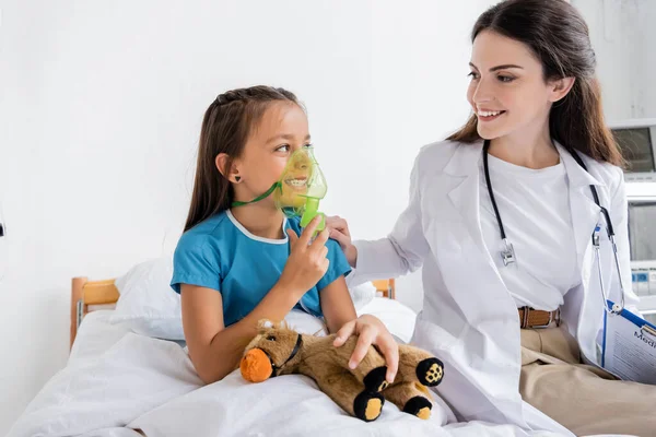 Médecin souriant avec presse-papiers regardant un enfant heureux avec masque à oxygène à l'hôpital — Photo de stock