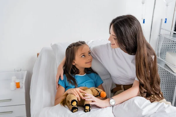 Sonriente madre abrazando hija con juguete suave en la cama en la clínica - foto de stock