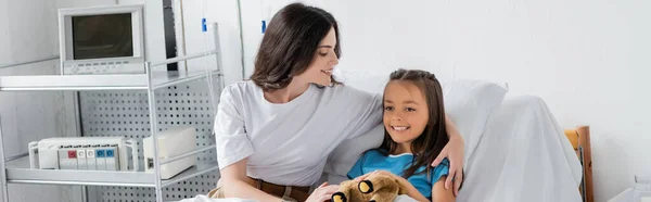 Positive woman hugging kid with toy on bed in hospital ward, banner — Stock Photo