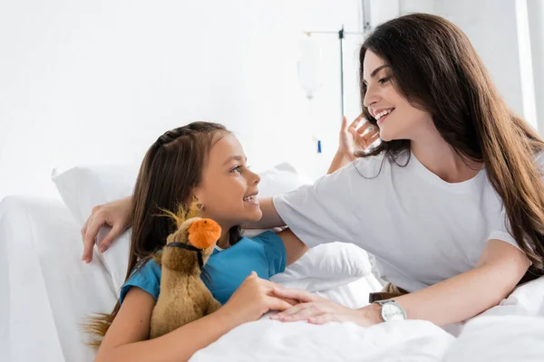 Positive kid hugging mother and holding soft toy on bed in clinic — Stock Photo