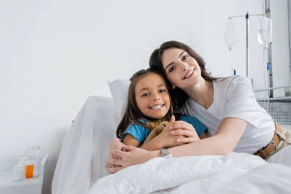 Positive woman and daughter looking at camera on bed in hospital ward — Stock Photo
