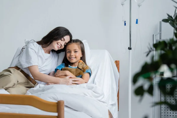 Sorrindo mulher abraçando filha com brinquedo na cama no hospital — Fotografia de Stock