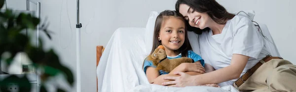 Joven madre abrazando al niño con juguete suave en la cama en la sala del hospital, pancarta - foto de stock