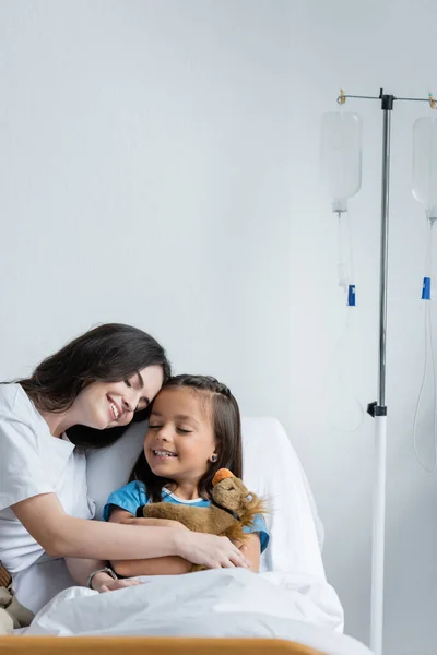 Parent touching hand of smiling child with soft toy on bed in clinic — Stock Photo