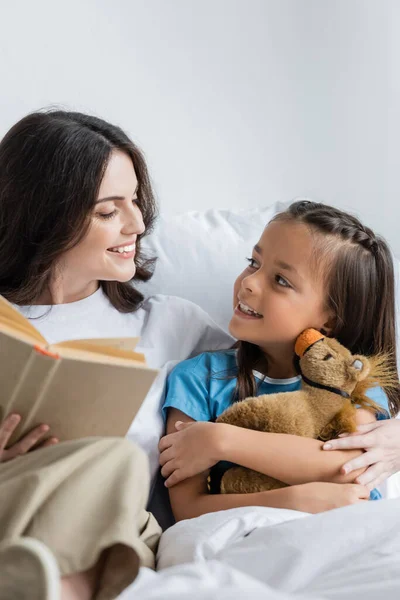 Mère tenant un livre près d'un enfant souriant avec jouet sur le lit de la clinique — Photo de stock