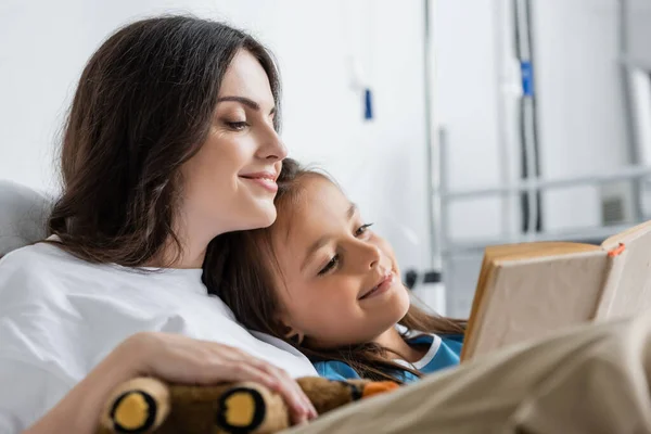Mujer positiva sosteniendo juguete suave mientras lee libro con la madre en la sala del hospital - foto de stock