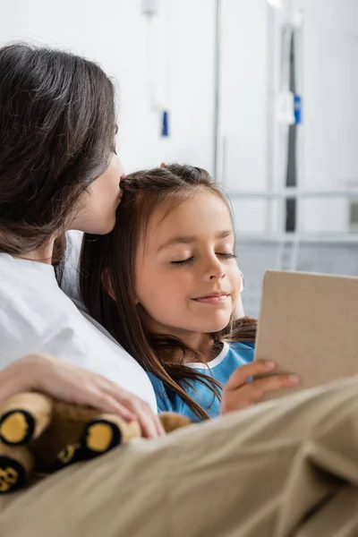 Mujer besando hija con libro borroso en la sala de hospital - foto de stock