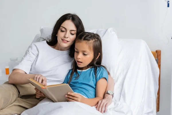 Mujer y niño leyendo libro juntos en la cama de la clínica - foto de stock
