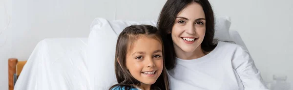 Позитивна жінка і дочка дивляться на камеру на лікарняному ліжку, банер — стокове фото