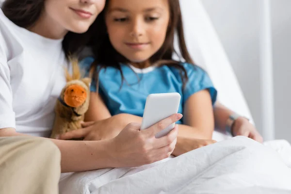 Mujer usando teléfono inteligente cerca de hija con juguete suave en la cama en el hospital - foto de stock