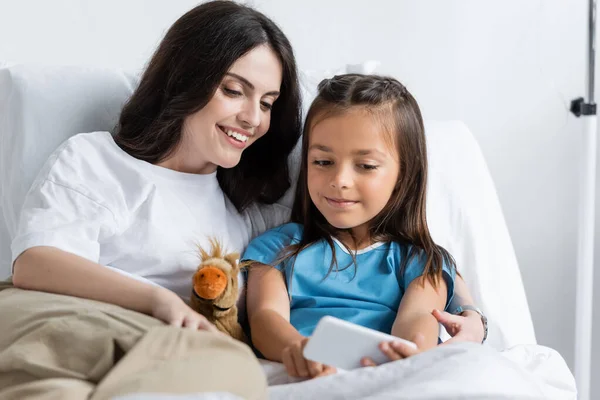 Sorridente madre abbracciando figlia con smartphone sul letto in clinica — Foto stock