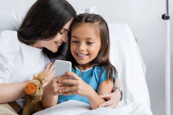 Chica alegre usando teléfono inteligente cerca de mamá y juguete suave en la sala de hospital - foto de stock