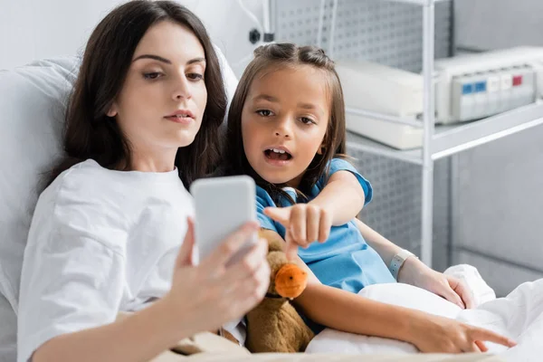 Ребенок указывает на смартфон рядом с мамой и мягкую игрушку на кровати в клинике — стоковое фото