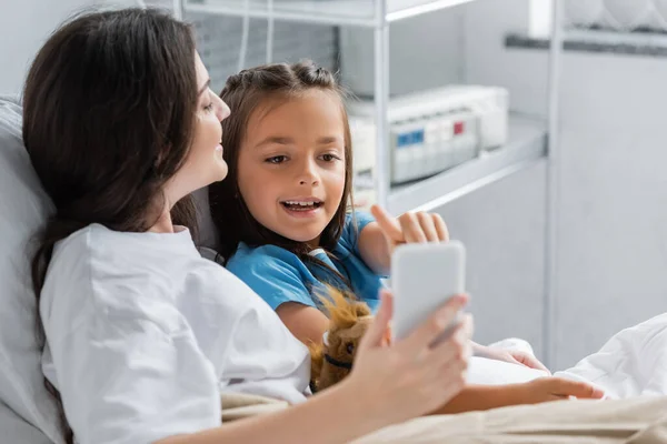 Menina sorridente apontando para o smartphone perto da mãe na cama na clínica — Fotografia de Stock