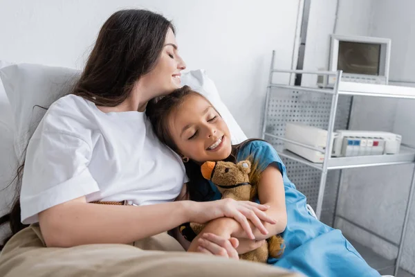 Веселая женщина держит за руку дочь в больничном халате на кровати в больнице — стоковое фото