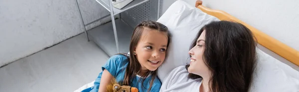 Vista de ángulo alto del niño sonriente en bata de paciente mirando a los padres en la cama del hospital, pancarta - foto de stock