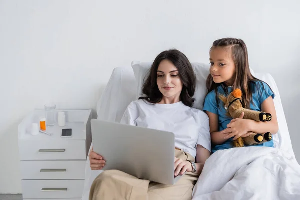 Madre sosteniendo portátil cerca de la hija en bata de paciente en la sala de hospital - foto de stock