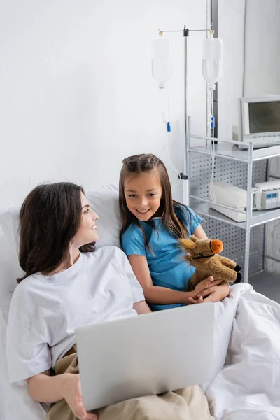 Усміхнена дитина в сукні пацієнта дивиться на ноутбук біля мами на лікарняному ліжку — стокове фото
