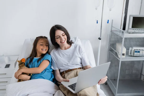 Mujer sosteniendo portátil cerca de niño en bata de paciente con juguete en la cama del hospital - foto de stock