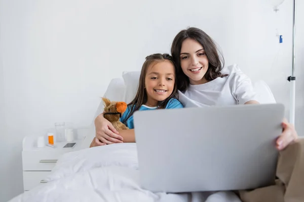 Genitore sorridente che tiene il computer portatile e abbraccia la figlia in abito paziente nel reparto ospedaliero — Foto stock