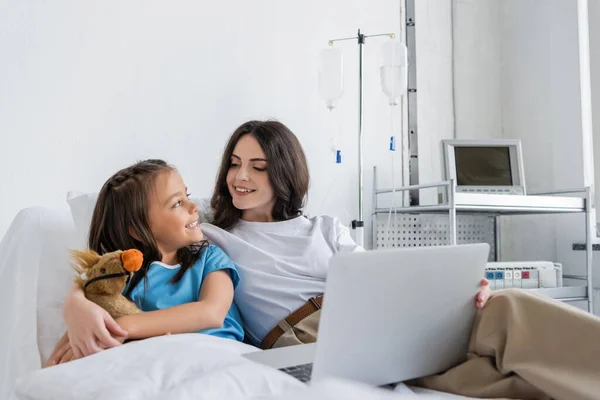 Niño en vestido de paciente sonriendo a la madre con el ordenador portátil en la cama en el hospital - foto de stock