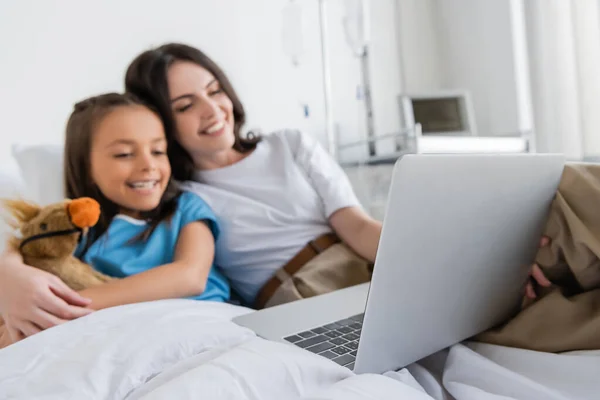 Laptop near blurred woman hugging daughter in patient gown on bed in clinic — Stock Photo