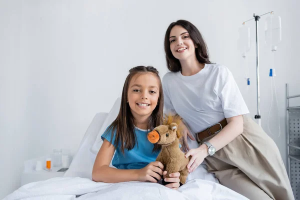 Улыбающийся ребенок в больничном платье с мягкой игрушкой рядом с матерью в больнице — стоковое фото
