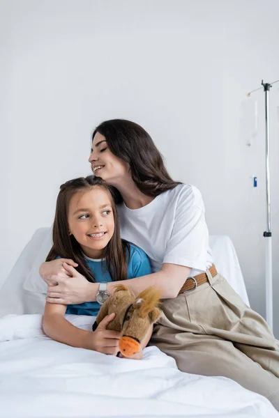 Madre abrazando hija en bata de paciente con juguete en la cama en la sala de hospital - foto de stock