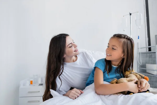Woman looking at kid in patient gown holding soft toy in hospital ward — Stock Photo
