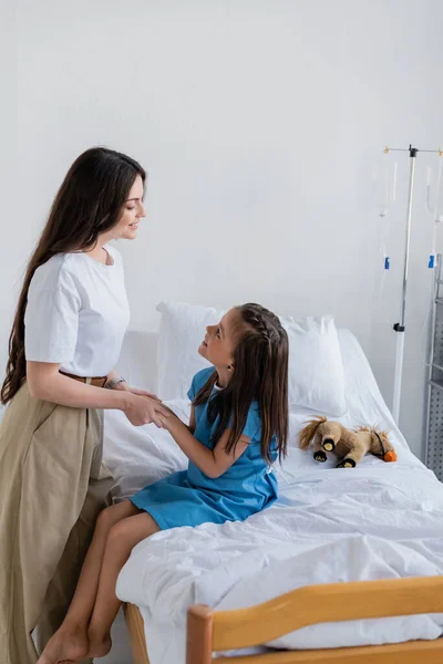 Вид сбоку мамы, держащей за руки дочь в больничном халате на кровати в клинике — стоковое фото