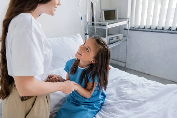 Donna che si tiene per mano la figlia in abito paziente sul letto in clinica — Foto stock
