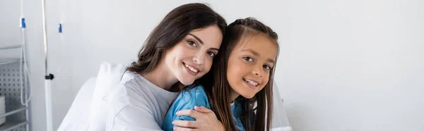 Mulher positiva abraçando filha e olhando para a câmera na clínica, banner — Fotografia de Stock