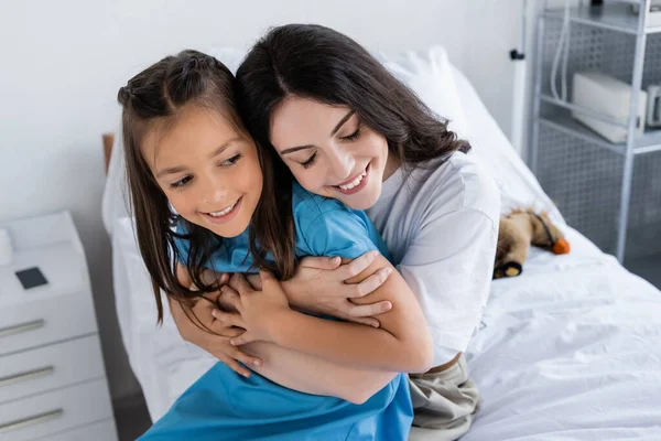 Lächelnde Mutter umarmt Tochter im Patientenkleid im Krankenhaus — Stockfoto