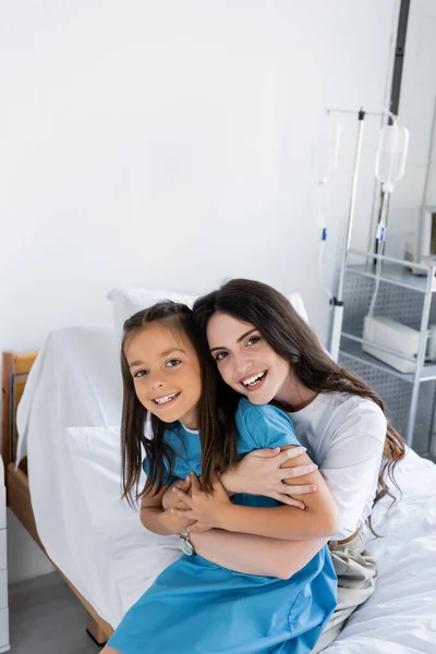 Позитивна жінка обіймає дочку в сукні пацієнта і дивиться на камеру в клініці — стокове фото