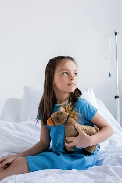 Niño en vestido de paciente sosteniendo juguete mientras está sentado en la cama en la clínica - foto de stock