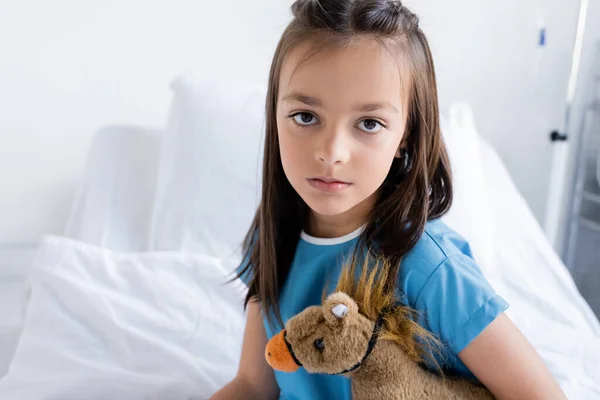 Enfant en robe de patient tenant un jouet et regardant une caméra sur un lit flou à l'hôpital — Photo de stock