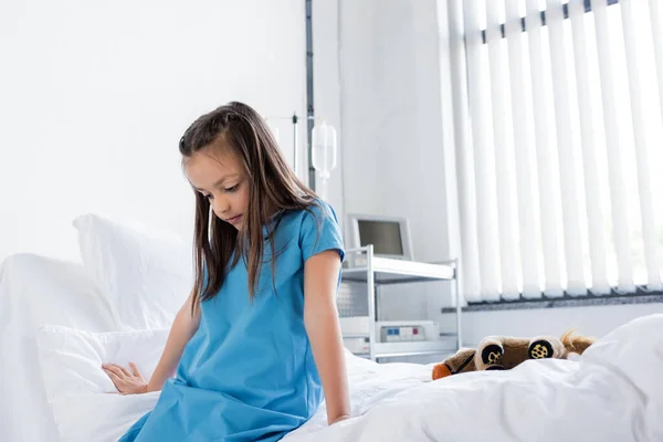 Menina em vestido de paciente sentado perto de brinquedo macio na cama no hospital — Fotografia de Stock